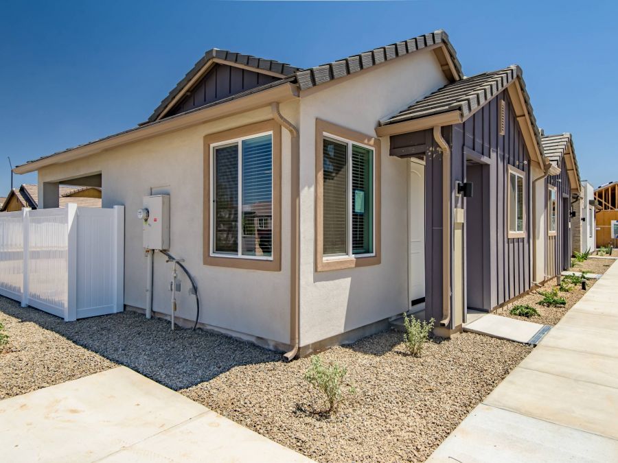 Single-story modern house with beige walls, gravel yard, white fence, and clear blue sky.